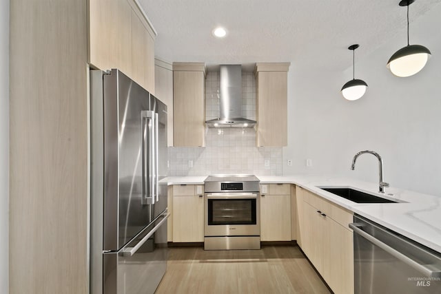 kitchen featuring wall chimney range hood, a sink, light wood finished floors, light brown cabinets, and stainless steel appliances