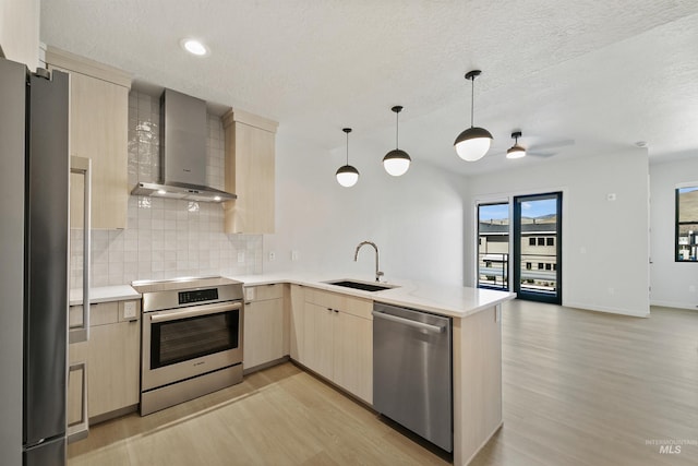 kitchen with wall chimney range hood, light brown cabinetry, appliances with stainless steel finishes, a sink, and a peninsula