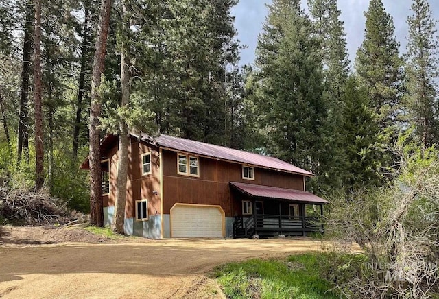 view of front facade featuring dirt driveway, metal roof, an attached garage, and a view of trees