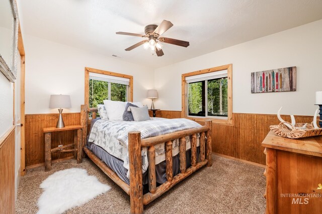 bedroom with a wainscoted wall, wooden walls, multiple windows, carpet flooring, and ceiling fan