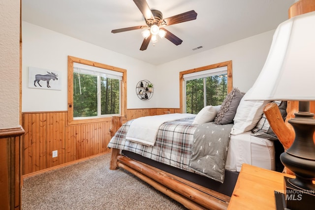 carpeted bedroom with wood walls, wainscoting, multiple windows, and a ceiling fan