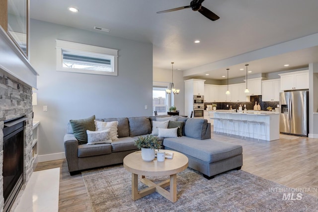 living room featuring a stone fireplace, a ceiling fan, light wood-style floors, a chandelier, and recessed lighting