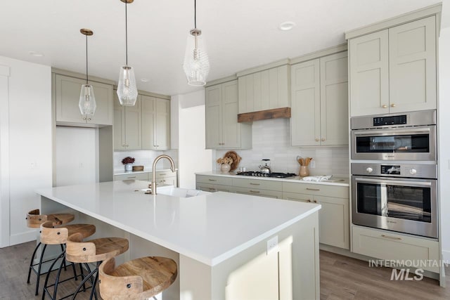 kitchen featuring stainless steel appliances, hanging light fixtures, sink, an island with sink, and backsplash