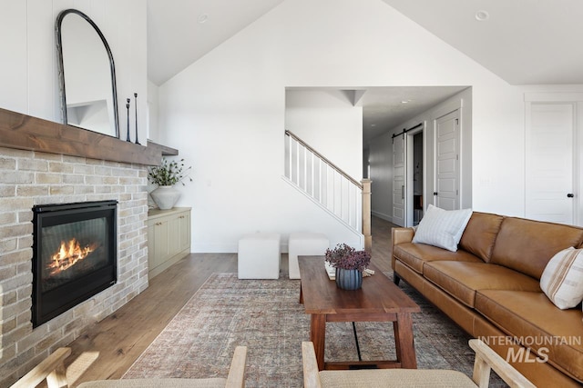 living room with a barn door, a brick fireplace, wood finished floors, high vaulted ceiling, and stairs