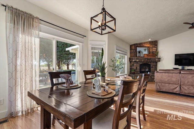 dining space with an inviting chandelier, light wood finished floors, a textured ceiling, a stone fireplace, and vaulted ceiling