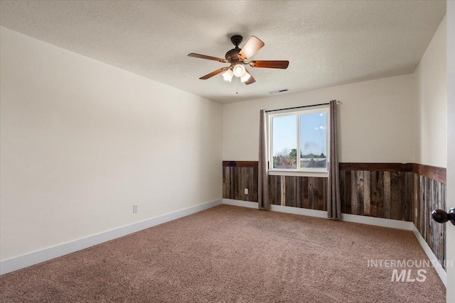 carpeted empty room with visible vents, wainscoting, ceiling fan, wooden walls, and a textured ceiling