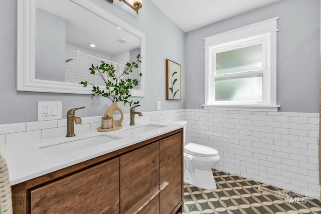 bathroom featuring tile walls, toilet, wainscoting, tiled shower, and double vanity