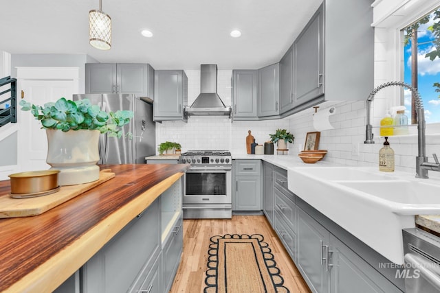 kitchen with gray cabinetry, appliances with stainless steel finishes, wall chimney exhaust hood, a sink, and wood counters