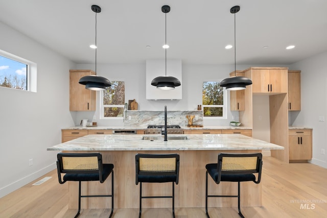 kitchen featuring a center island with sink, decorative light fixtures, light brown cabinetry, and light stone counters