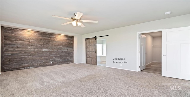 unfurnished bedroom featuring carpet flooring, a barn door, baseboards, ceiling fan, and wooden walls