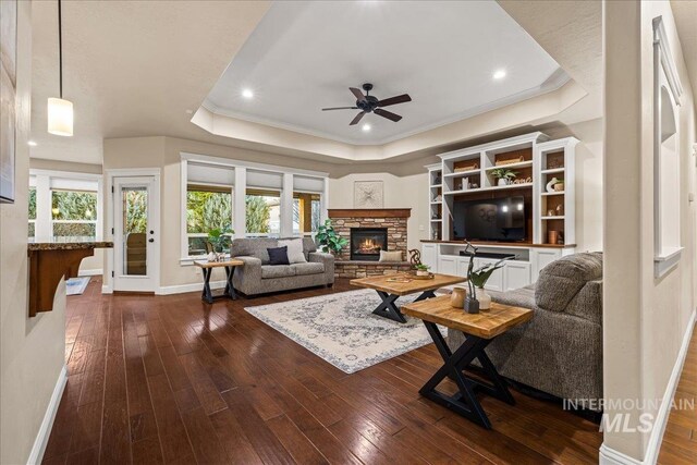 living area with a raised ceiling, plenty of natural light, a stone fireplace, and dark wood-style floors