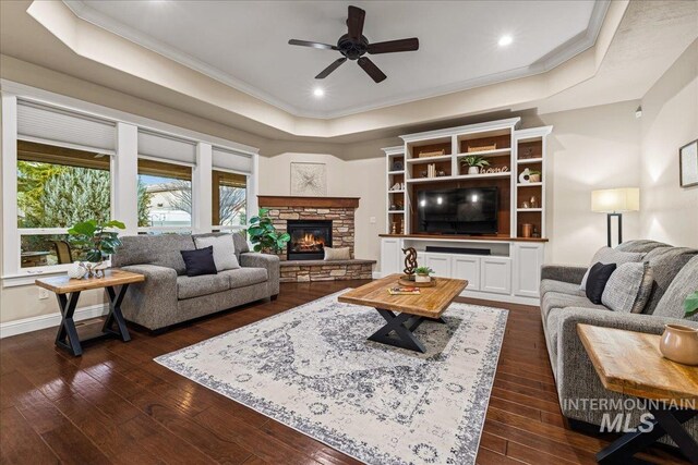 living room with baseboards, dark wood-style flooring, a stone fireplace, crown molding, and a raised ceiling