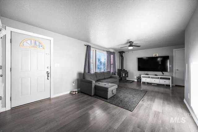 living room featuring baseboards, dark wood finished floors, a ceiling fan, a wood stove, and a textured ceiling