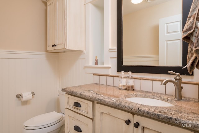 half bathroom featuring vanity, a wainscoted wall, a decorative wall, and toilet