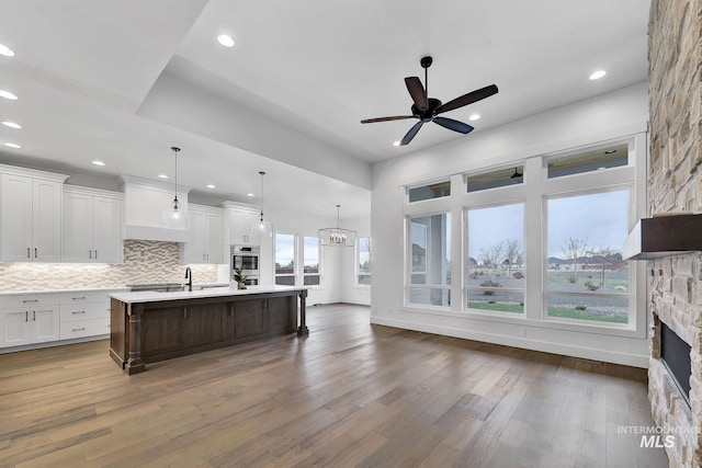 kitchen with white cabinetry, an island with sink, dark wood-type flooring, a fireplace, and ceiling fan with notable chandelier