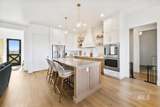 kitchen featuring recessed lighting, premium range hood, open shelves, a center island with sink, and appliances with stainless steel finishes