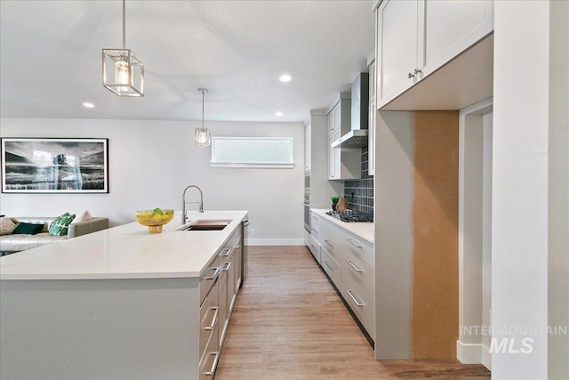 kitchen featuring sink, wall chimney exhaust hood, tasteful backsplash, decorative light fixtures, and a center island with sink