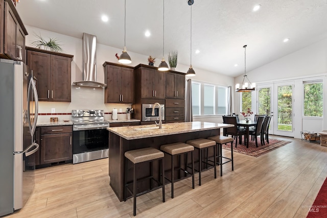 kitchen featuring wall chimney exhaust hood, an island with sink, pendant lighting, stainless steel appliances, and light stone countertops