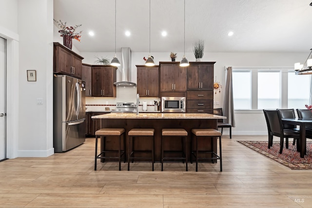 kitchen with appliances with stainless steel finishes, hanging light fixtures, light stone counters, a center island with sink, and wall chimney exhaust hood