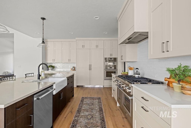 kitchen with custom exhaust hood, dark brown cabinetry, an island with sink, white cabinetry, and stainless steel appliances