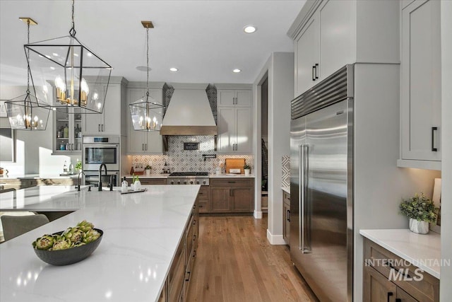 kitchen featuring custom exhaust hood, hanging light fixtures, appliances with stainless steel finishes, glass insert cabinets, and light stone countertops