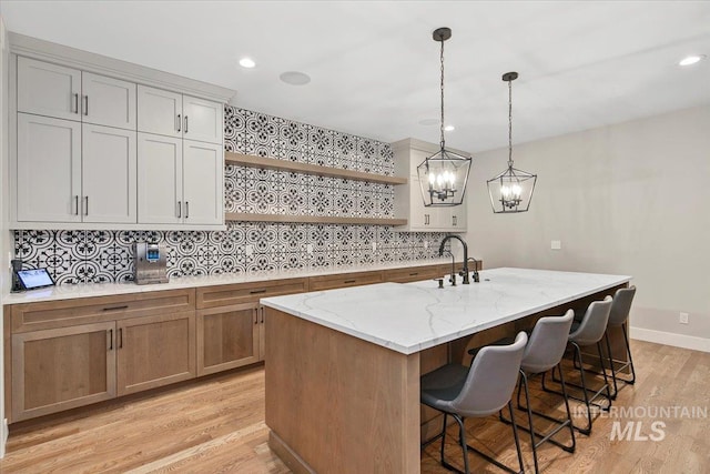 kitchen with light stone counters, a kitchen island with sink, hanging light fixtures, decorative backsplash, and open shelves