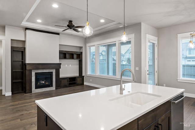 kitchen with ceiling fan, sink, hanging light fixtures, a tray ceiling, and a kitchen island with sink