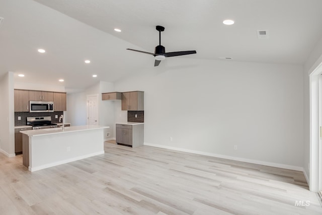 kitchen with vaulted ceiling, light wood-type flooring, appliances with stainless steel finishes, an island with sink, and decorative backsplash