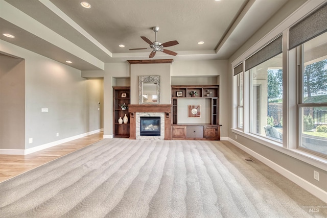unfurnished living room with a raised ceiling, light carpet, baseboards, a glass covered fireplace, and ceiling fan