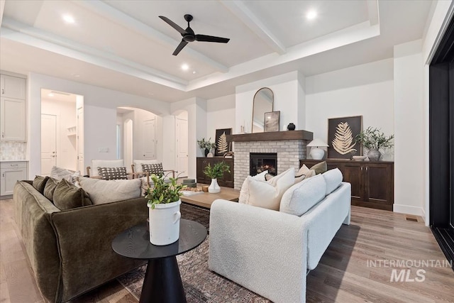 living room featuring a brick fireplace, light wood-type flooring, beamed ceiling, ceiling fan, and a raised ceiling