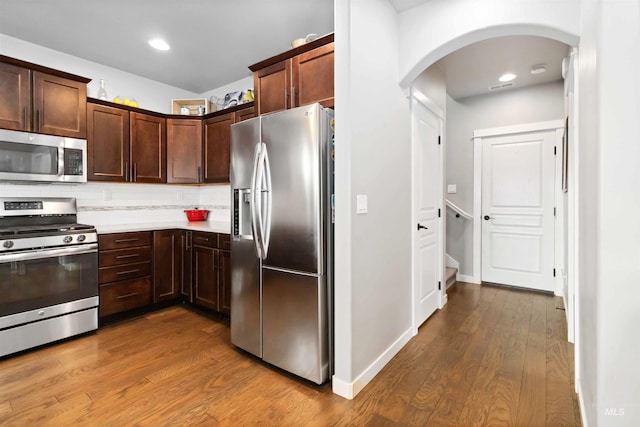 kitchen with dark brown cabinetry, stainless steel appliances, wood finished floors, light countertops, and backsplash