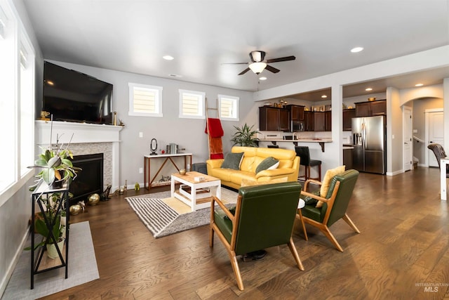 living area featuring arched walkways, a tiled fireplace, dark wood-type flooring, ceiling fan, and baseboards