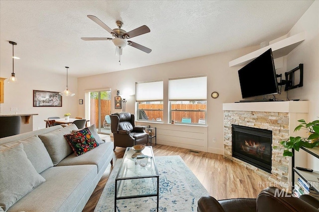 living area featuring a textured ceiling, a stone fireplace, wood finished floors, and a ceiling fan