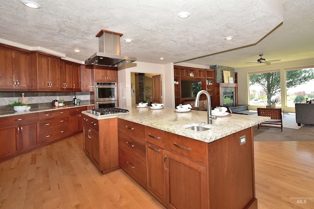 kitchen with a sink, a ceiling fan, island exhaust hood, light wood-style flooring, and stainless steel double oven