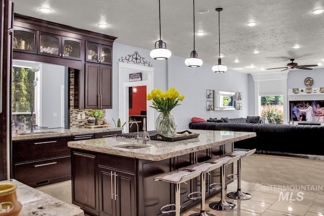 kitchen featuring pendant lighting, sink, dark brown cabinetry, light stone counters, and a center island with sink