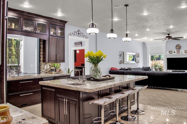 kitchen featuring sink, decorative light fixtures, dark brown cabinets, an island with sink, and light stone countertops