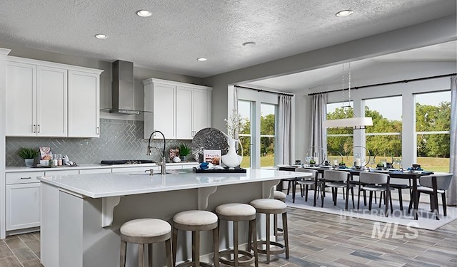 kitchen featuring white cabinetry, wall chimney exhaust hood, backsplash, an island with sink, and lofted ceiling