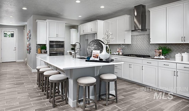 kitchen with a center island with sink, white cabinetry, wall chimney range hood, and appliances with stainless steel finishes