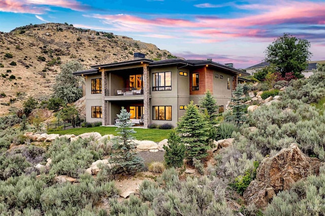 back of house at dusk featuring a chimney, stucco siding, a mountain view, a balcony, and stone siding