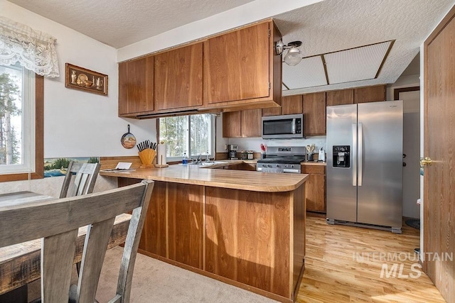 kitchen with light wood-type flooring, appliances with stainless steel finishes, a peninsula, brown cabinetry, and light countertops