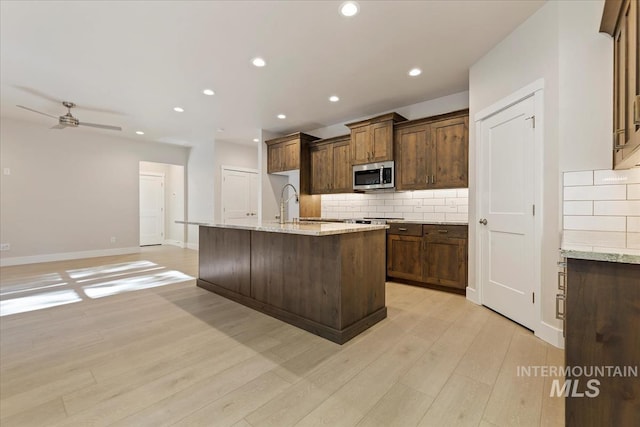 kitchen with ceiling fan, light hardwood / wood-style floors, light stone countertops, and an island with sink