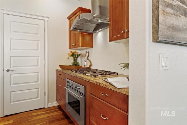 kitchen featuring stainless steel appliances, wood finished floors, wall chimney exhaust hood, and brown cabinetry