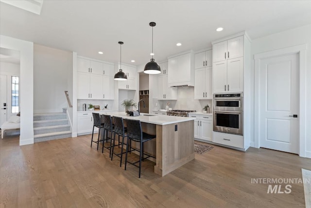 kitchen with double oven, hanging light fixtures, sink, white cabinets, and a kitchen island with sink