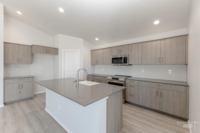 kitchen with vaulted ceiling, backsplash, stainless steel appliances, a kitchen island with sink, and light wood-type flooring