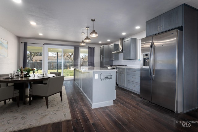 kitchen with light stone counters, wall chimney exhaust hood, pendant lighting, an island with sink, and appliances with stainless steel finishes