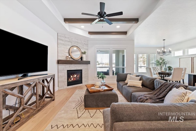 living area featuring ceiling fan with notable chandelier, a stone fireplace, light wood-style floors, and a raised ceiling