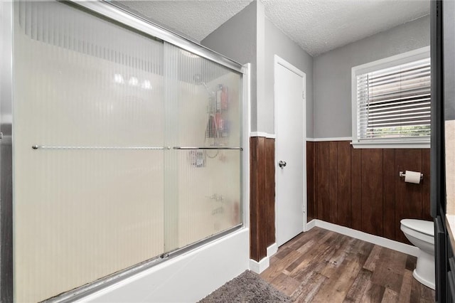 bathroom with bath / shower combo with glass door, a textured ceiling, wooden walls, hardwood / wood-style floors, and toilet