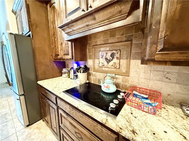 kitchen with stainless steel refrigerator, tasteful backsplash, light stone countertops, custom range hood, and black electric cooktop