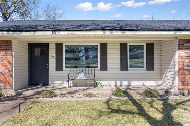 view of exterior entry with brick siding, a porch, and roof with shingles