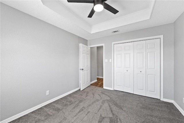 unfurnished bedroom with a raised ceiling, ceiling fan, a closet, and dark colored carpet